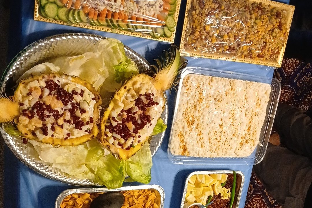 A spread of home-cooked dishes laid out on a long table, including pineapple fruit chaat, kheer, corn chaat, salad platters, pasta and rice trays